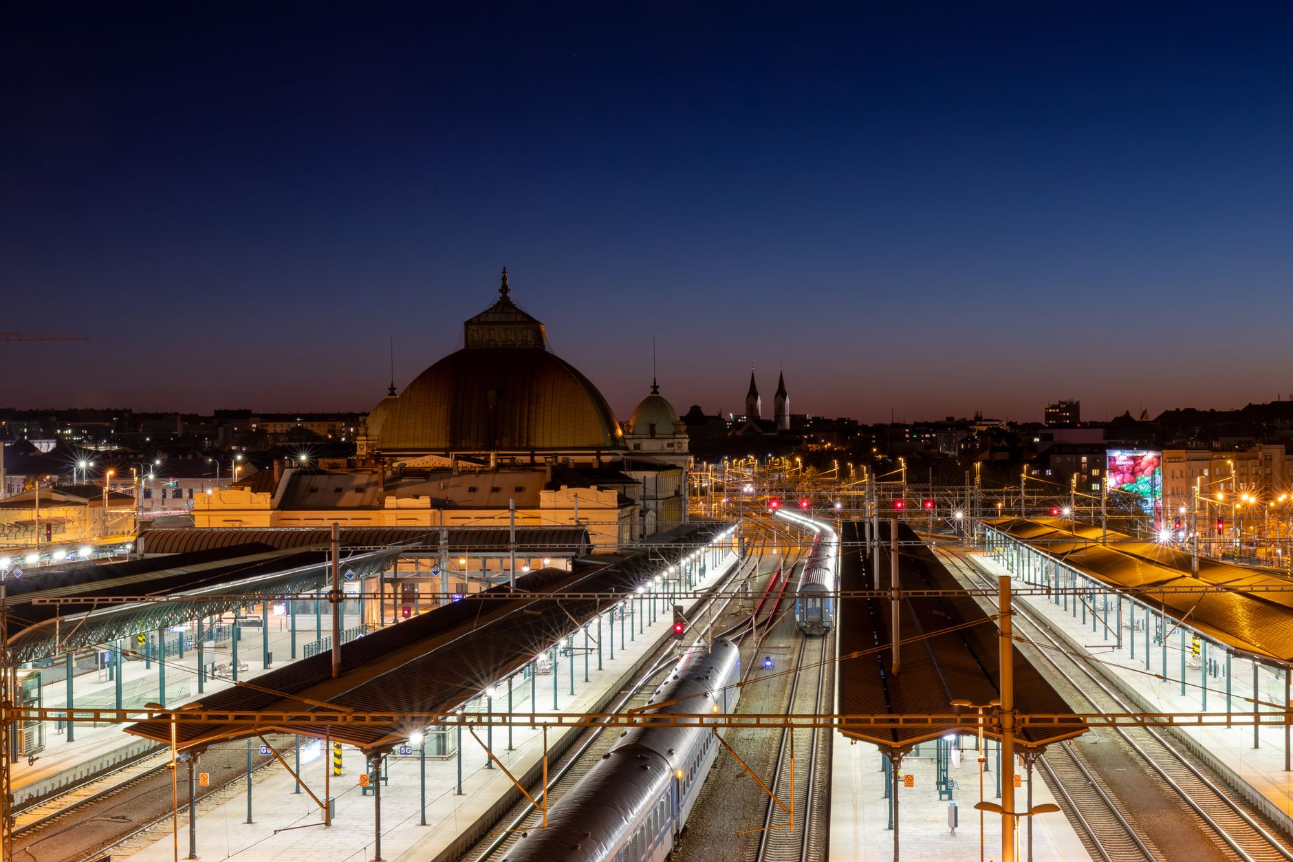 Lit platforms, tracks and overhead wires at a large railway station at dusk, with a domed station building and city rooftops behind