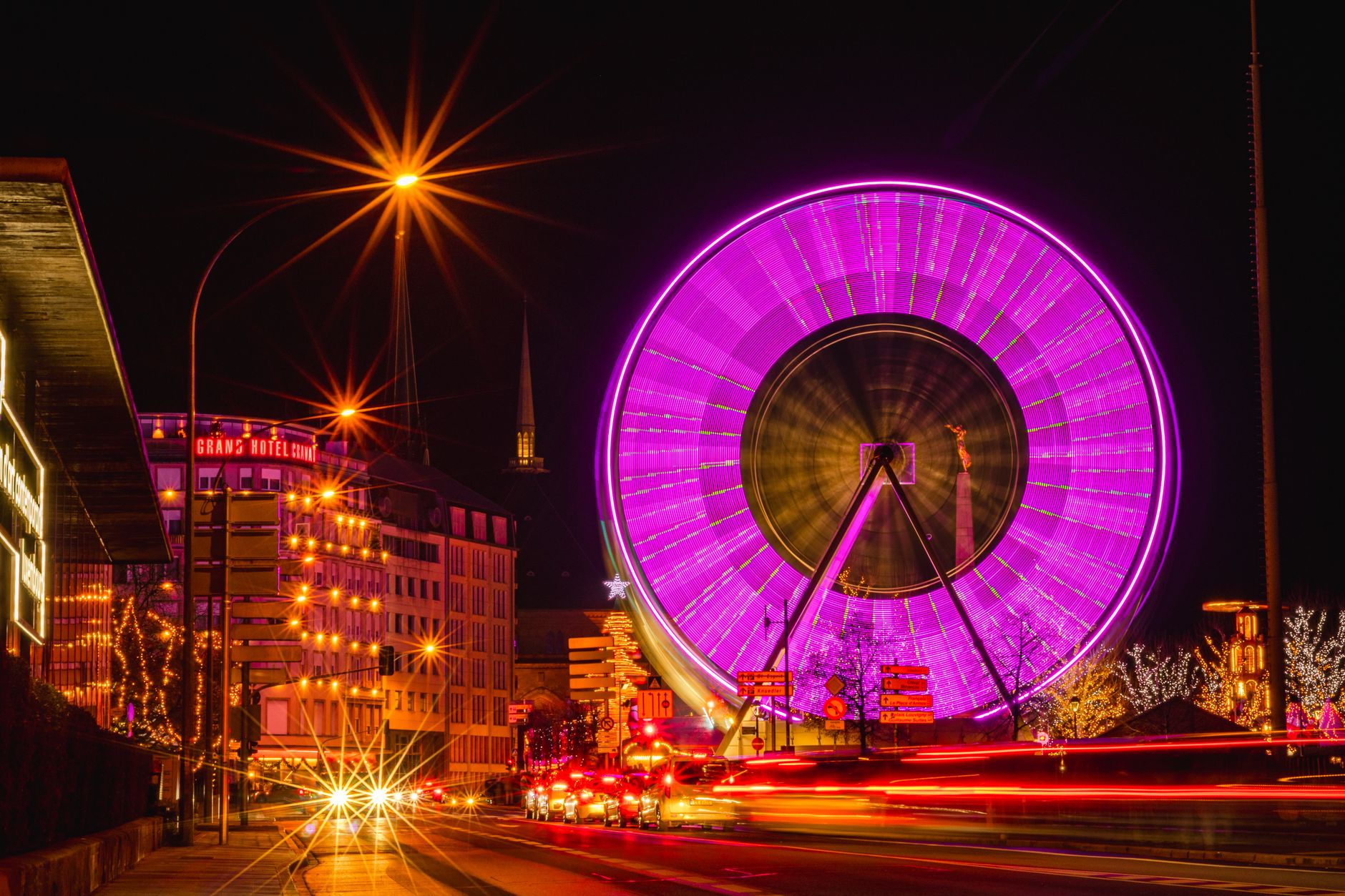 A ferris wheel lit in magenta turning at night above a city street, with fairy lights in the trees and traffic light trails